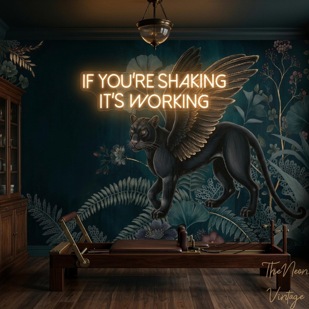 A room with a dark green wall mural featuring a winged black panther and botanical elements, a Pilates reformer, and a neon sign that reads "IF YOU'RE SHAKING IT'S WORKING."