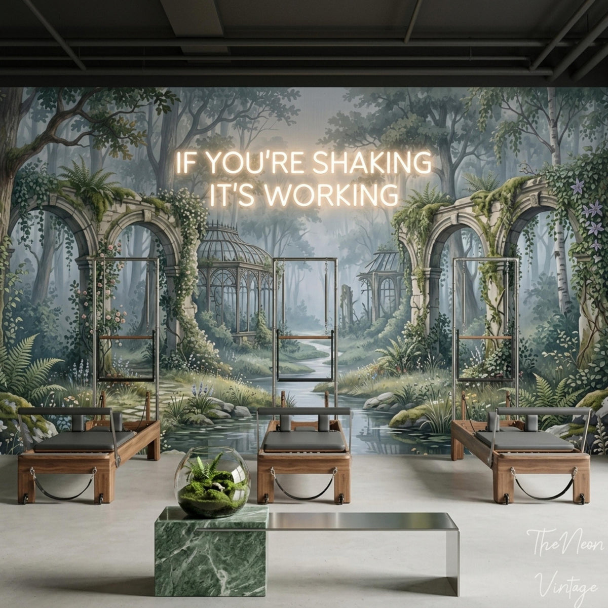 A fitness studio with three Pilates reformers and a neon sign reading "IF YOU'RE SHAKING IT'S WORKING" on a wall mural of a forest with ruins.