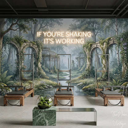 A fitness studio with three Pilates reformers and a neon sign reading "IF YOU'RE SHAKING IT'S WORKING" on a wall mural of a forest with ruins.