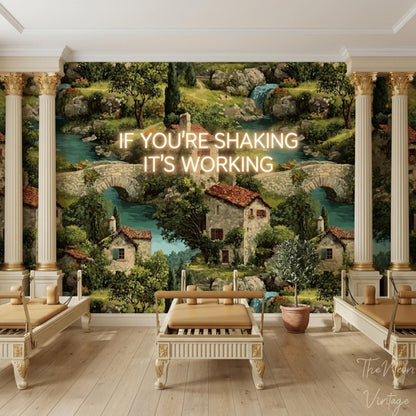 A room with two white benches and columns, against a wallpaper featuring a painted village scene with houses, trees, and a river, with the text "IF YOU'RE SHAKING IT'S WORKING" in neon letters.