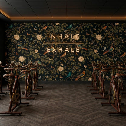 A fitness room with dark floral wallpaper features rows of stationary exercise bikes and a neon sign that reads inhale exhale.