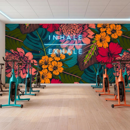 An indoor cycling studio has a dark tropical floral accent wall with neon text reading Inhale Exhale above several exercise bikes.