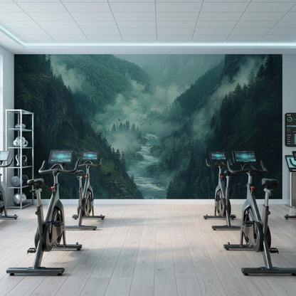 Several stationary exercise bikes face a large wall mural depicting a misty mountain river valley in a bright gym.