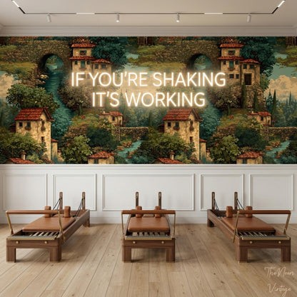 A room with wooden flooring features a large green and brown mural of an Italian village on the wall, overlaid with a glowing neon sign reading If You Are Shaking It Is Working, and three Pilates reformers in front.