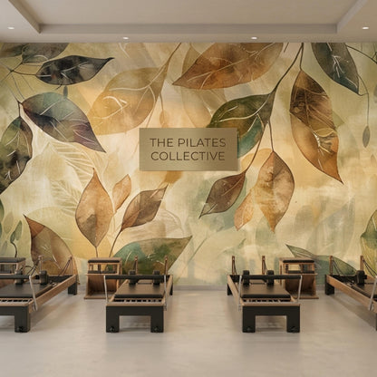 A fitness space displays four black pilates reformers on a concrete floor in front of a botanical watercolor leaf wallpaper mural with a sign that reads The Pilates Collective.