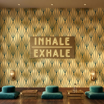 A meditation space features low teal floor cushions and small gold tables beneath a glowing neon sign that reads inhale exhale on gold and dark green art deco geometric wallpaper.