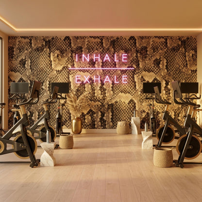 A gym room with exercise bikes features a brown snake skin wall mural illuminated by a pink neon sign that reads inhale exhale.
