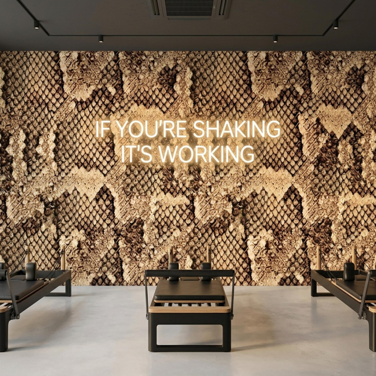 A pilates studio with reformer machines features a brown snake skin wall mural with a white neon sign that reads if you are shaking it is working.