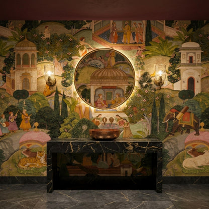 A round illuminated mirror hangs on a Mughal miniature art wallpaper wall above a dark marble console table and copper bowl.