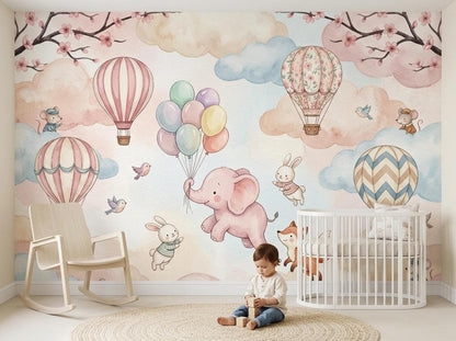 A child sits on a round rug in front of a watercolor nursery wall mural featuring animals, hot air balloons, and pastel clouds.
