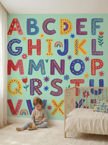 A toddler sits on the floor playing with a wooden toy against a mint green wall featuring a folk art alphabet mural.
