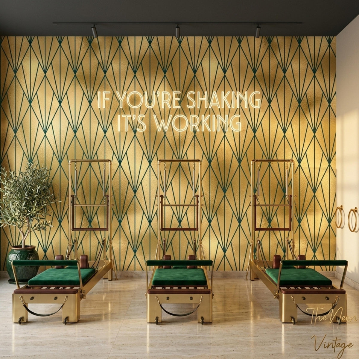 A gym room features gold and dark green geometric art deco wallpaper on the back wall, topped with a glowing neon sign that reads IF YOU'RE SHAKING IT'S WORKING above three pilates reformers with green cushions.