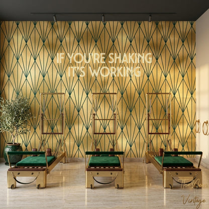 A gym room features gold and dark green geometric art deco wallpaper on the back wall, topped with a glowing neon sign that reads IF YOU'RE SHAKING IT'S WORKING above three pilates reformers with green cushions.