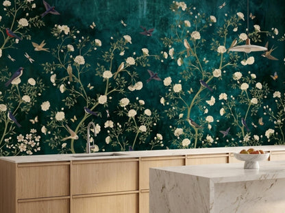 A kitchen counter and marble island sit in front of a moody teal botanical wallpaper mural with white blossoms and birds.