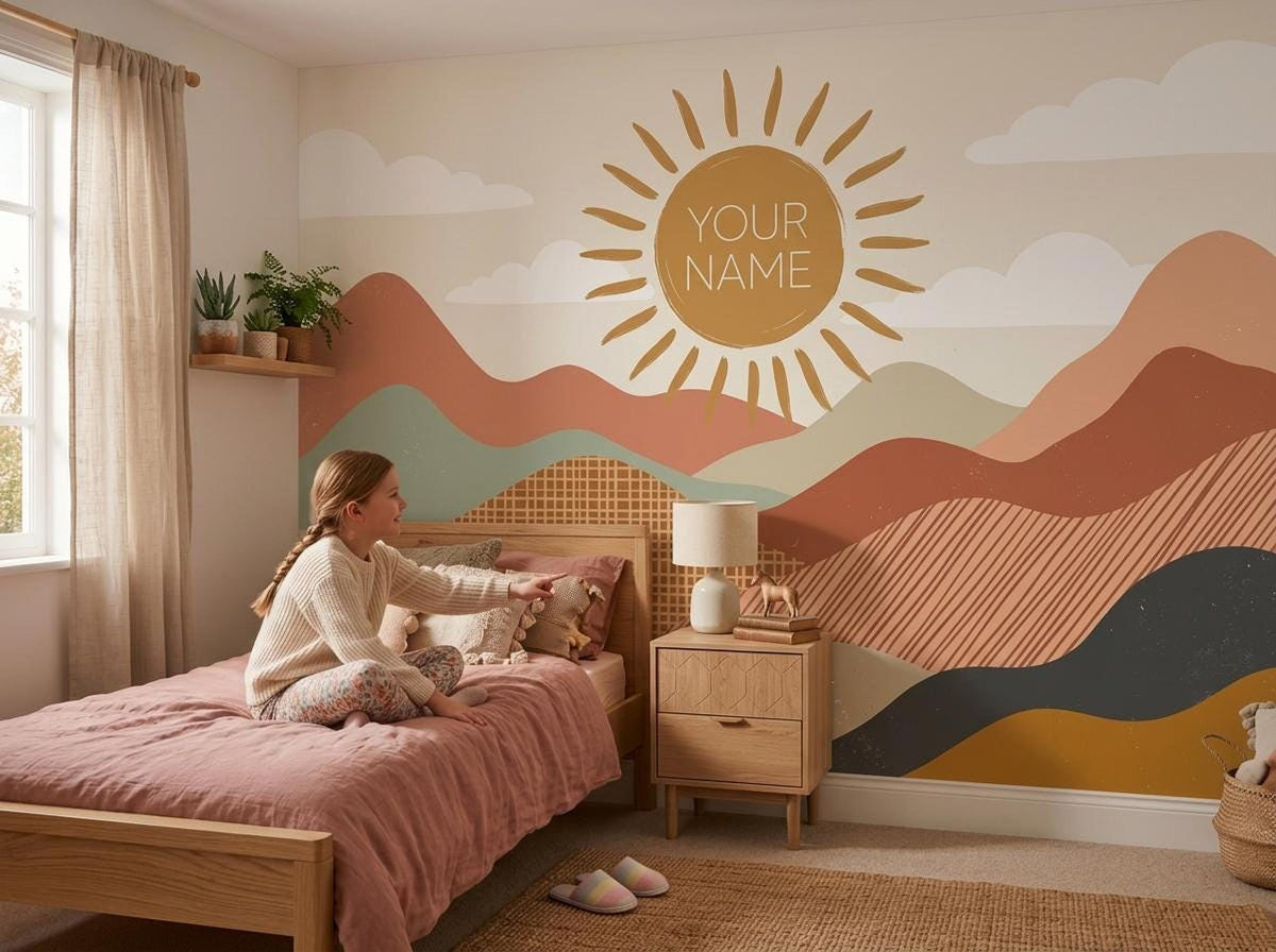 A child sits on a wooden bed in a bedroom decorated with a boho sun and mountain wall mural displaying the text YOUR NAME.