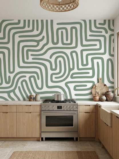 A kitchen backsplash featuring sage green maze pattern wallpaper above wooden cabinets and a stainless steel stove.