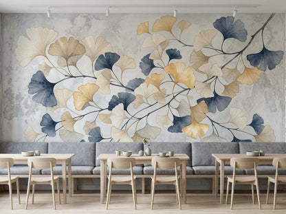 A row of wooden dining tables and gray upholstered bench seating lines a textured concrete wall covered in a botanical mural of gold and blue ginkgo leaves.