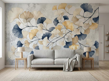 A living room features a large ginkgo leaf wall mural in shades of gold, beige, and navy blue over a gray textured background, with a gray sofa and wooden accent tables.