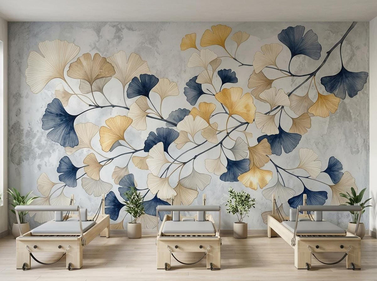 Three pilates reformer machines are positioned in front of a gray and white textured wall mural featuring large botanical ginkgo leaves in gold, beige, and dark blue.