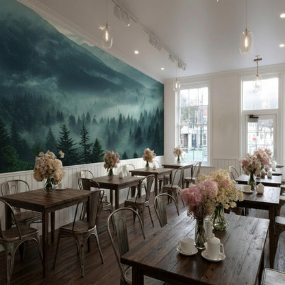 A bright cafe interior has wooden tables decorated with flower vases, placed next to a wall mural of misty mountains and pine trees.