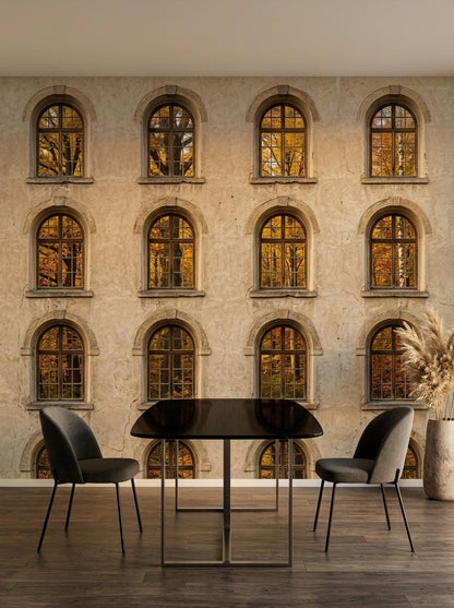 A dining space with a dark rectangular table and two dark gray chairs set in front of a textured wall mural of an antique stone building facade with autumn window views.