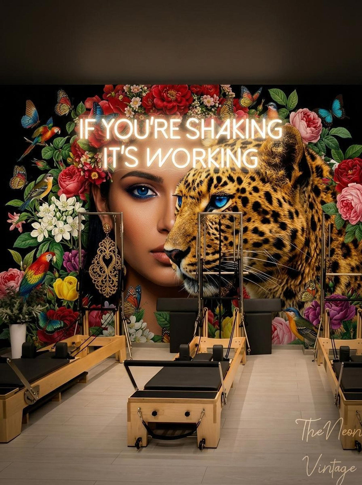 A pilates studio featuring the floral leopard mural on the back wall with a neon sign reading If You're Shaking It's Working above pilates reformers.