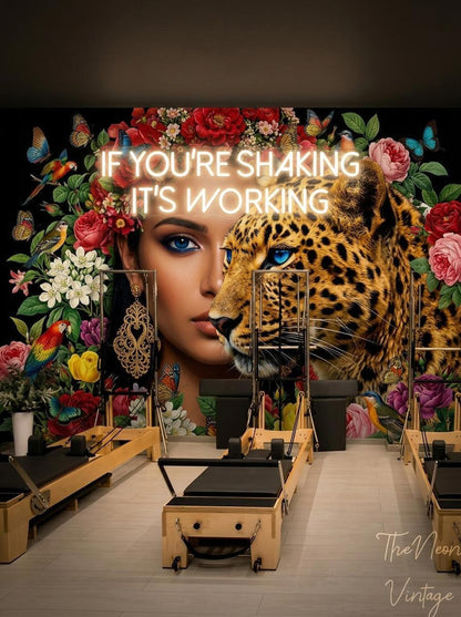 A pilates studio featuring the floral leopard mural on the back wall with a neon sign reading If You're Shaking It's Working above pilates reformers.