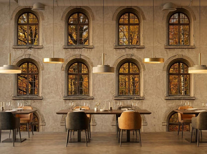 A restaurant interior with wooden dining tables and chairs against a large wall mural depicting an old stone building facade with arched windows looking out at autumn trees.