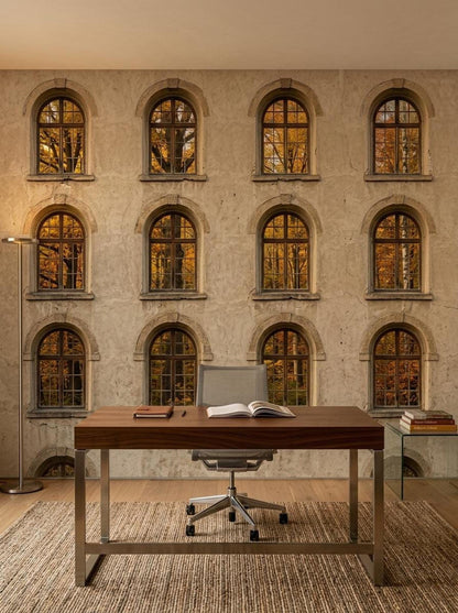 A home office setup with a wooden desk, office chair, and rug against a wallpaper mural of an old Italian stone building facade with arched autumn windows.
