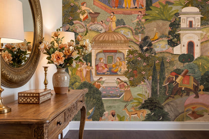 A detailed Mughal garden mural wallpaper featuring figures, animals, and architecture, shown on a wall behind a wooden console table with decor.